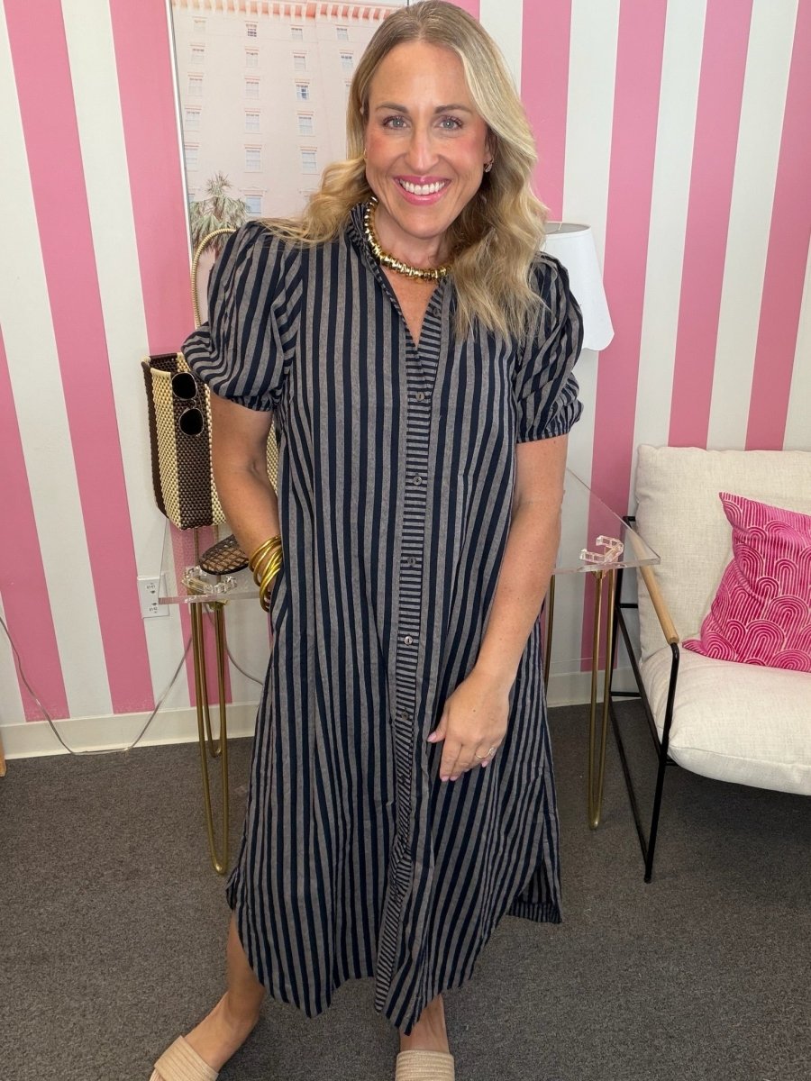 A woman wearing a black and white vertically striped midi dress with puff sleeves and a button-down front, standing in front of a pink and white striped wall.