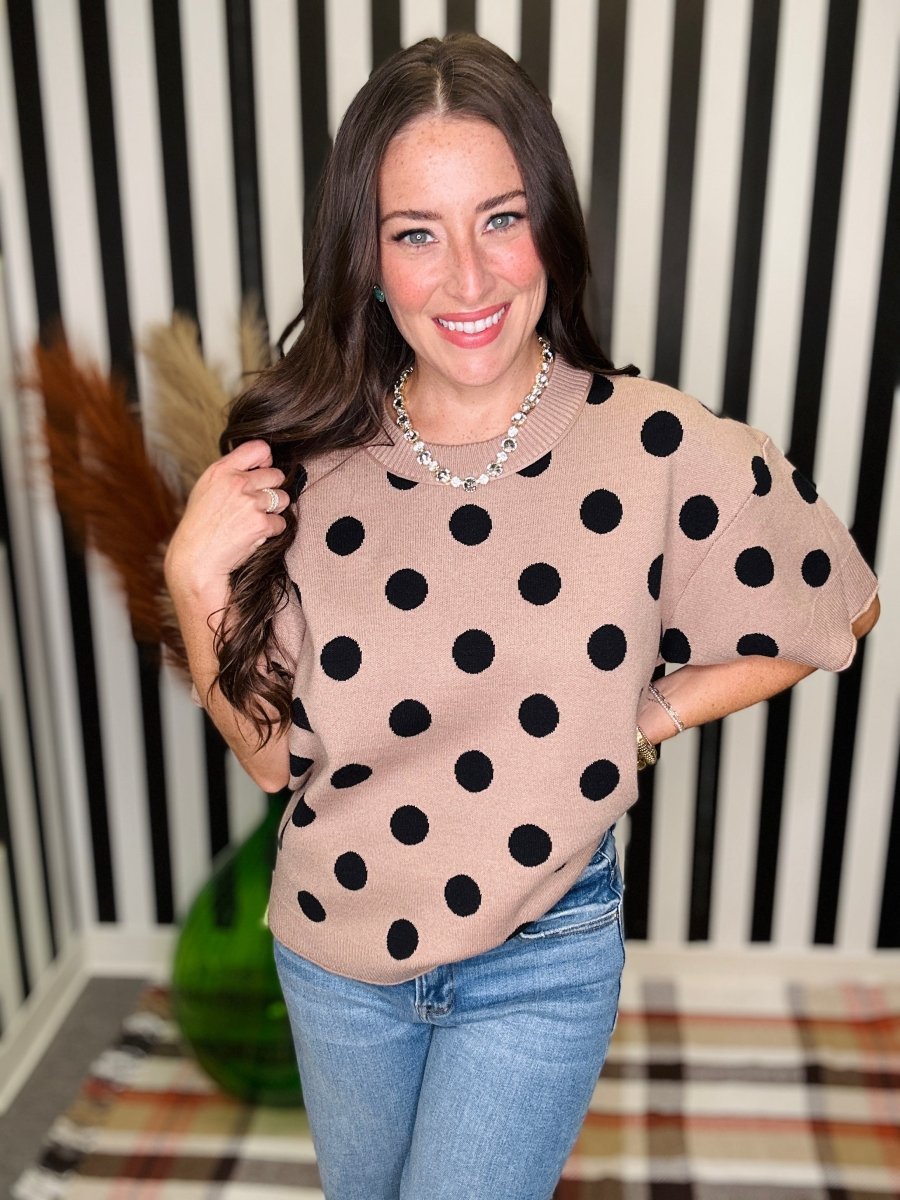 A woman wearing a beige knit sweater with large black polka dots and light blue jeans, standing in front of a black and white striped wall.