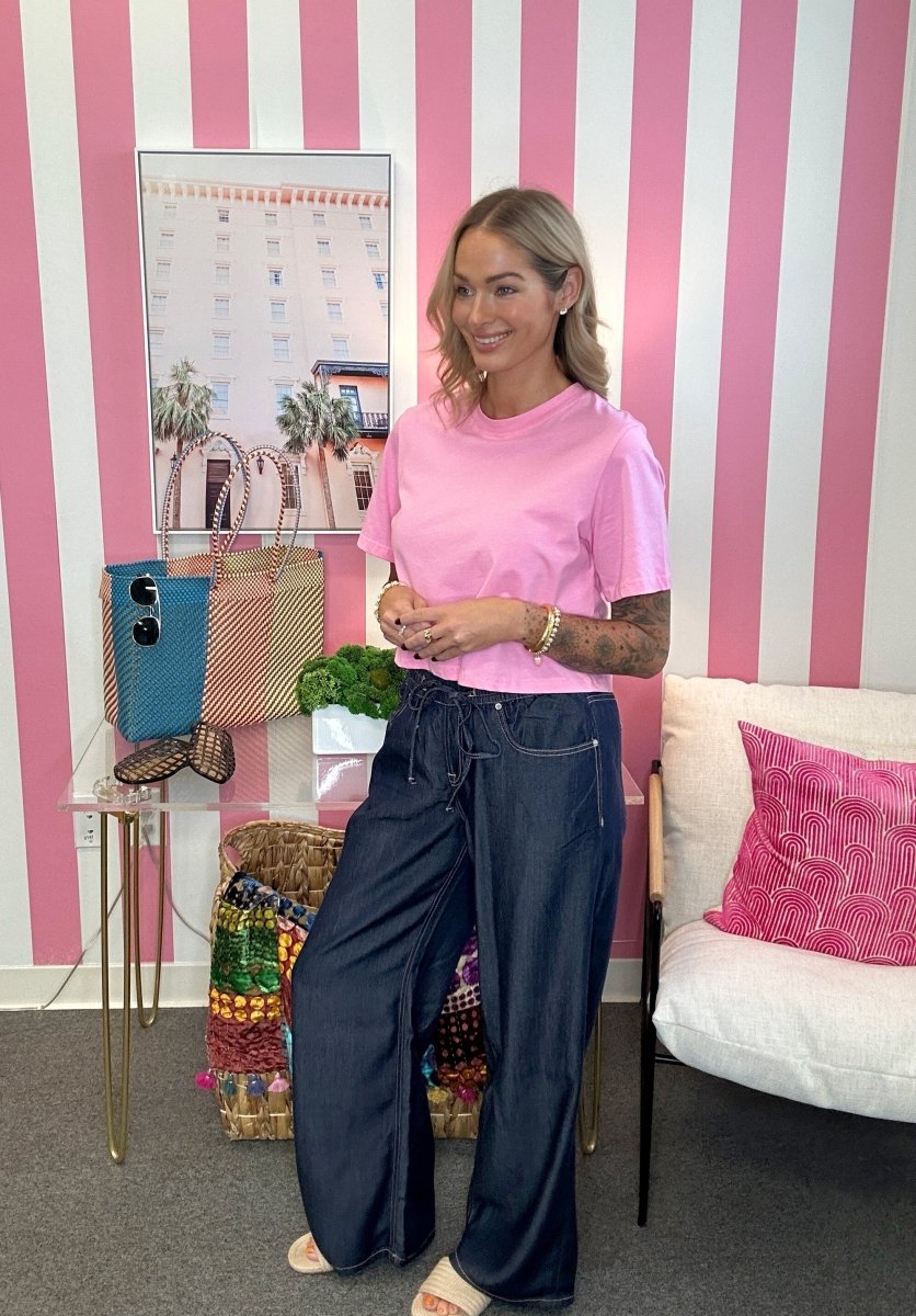 A woman stands in a pink and white striped room wearing a light pink short-sleeve t-shirt and dark navy wide-leg jeans. She is posing next to a white sofa with a pink pillow and a small table holding a blue bag and a woven tote.