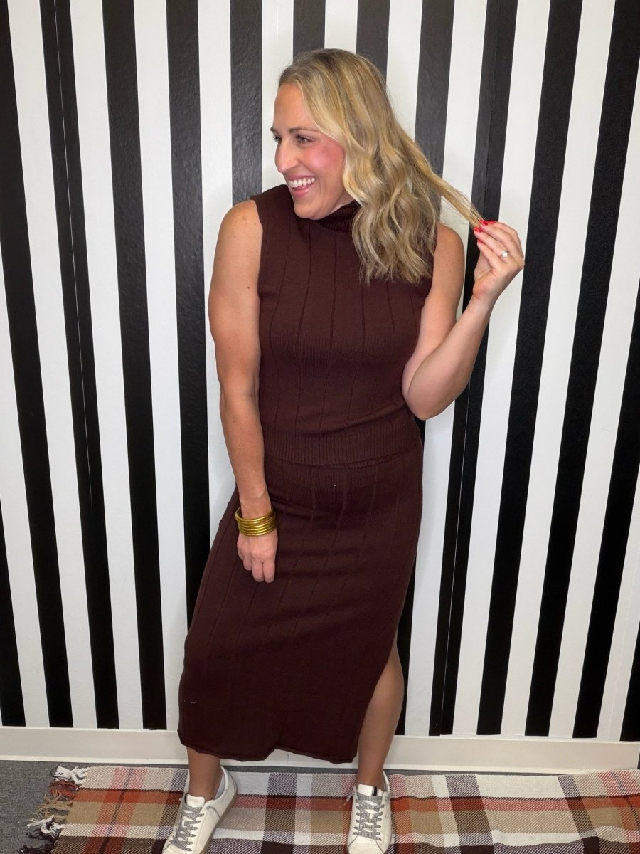 A woman modeling a sleeveless, ribbed brown mock-neck midi dress with a side slit, standing in front of a black and white striped wall.