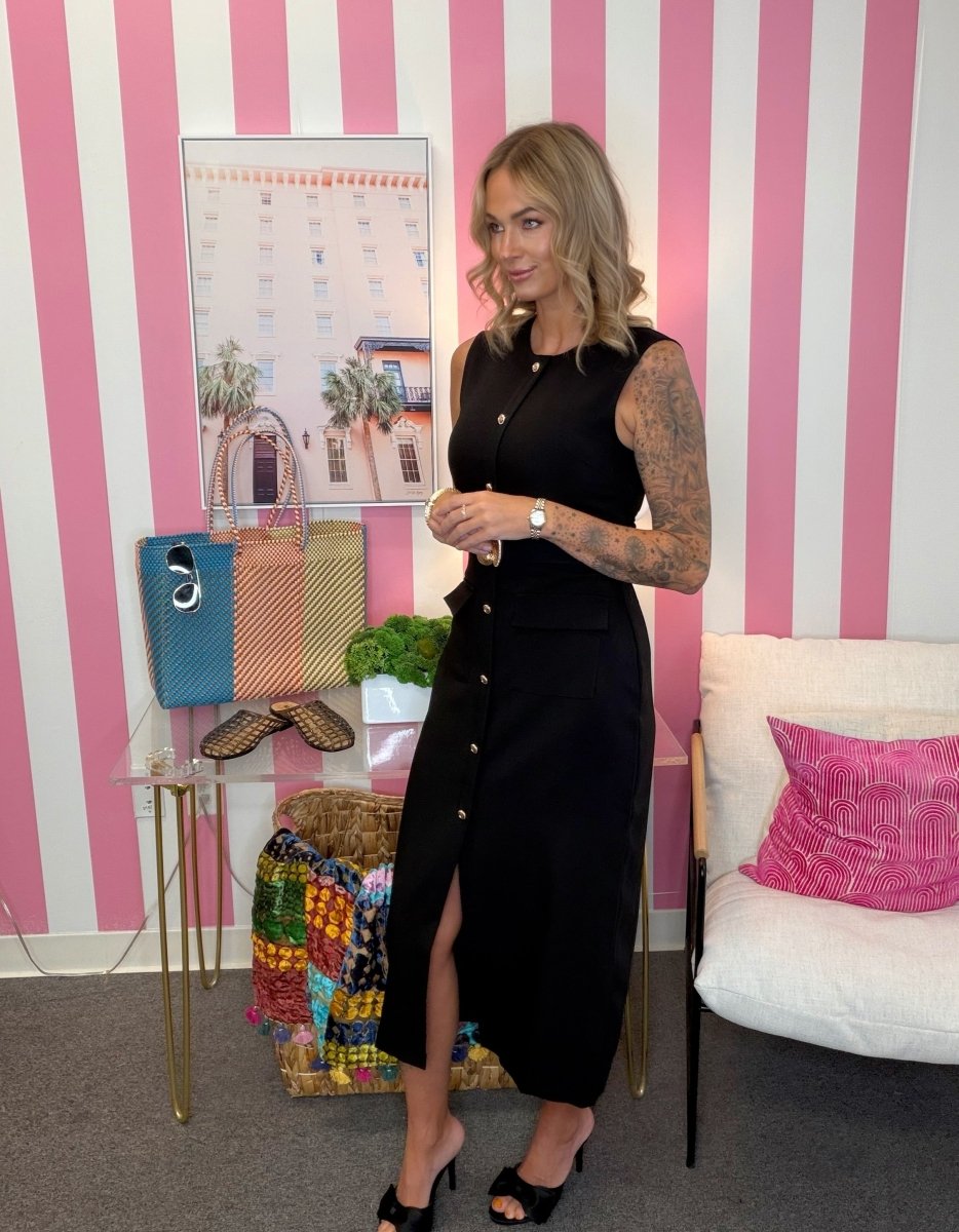 A woman with arm tattoos stands in a room with pink and white striped wallpaper, wearing a sleeveless black midi dress with a high slit and front buttons. She is holding a small cup next to a glass table with handbags and shoes, with a white armchair and pink pillow in the background.