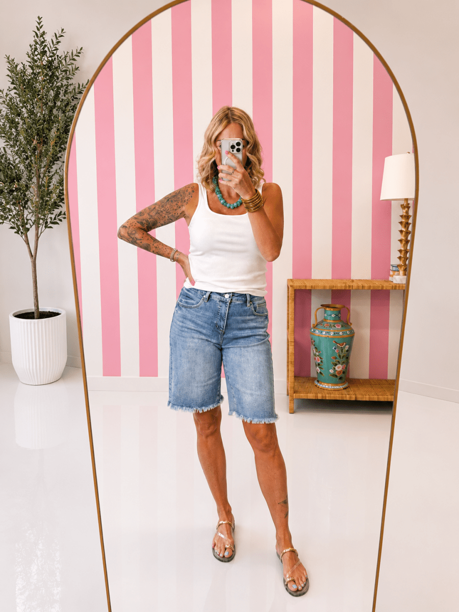 Woman taking a selfie in front of a pink and white striped wall with a plant and shelf in the background.