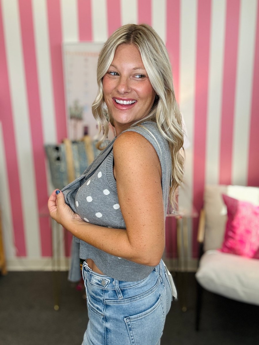 A smiling blonde woman wearing a gray sleeveless knit top with white polka dots and light blue jeans, standing in front of a pink and white striped wall.