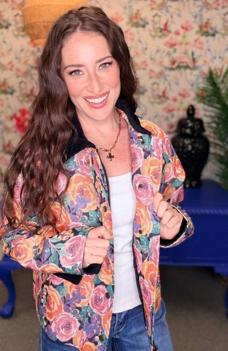 A woman smiling and wearing a colorful floral bomber jacket with a white top and blue jeans, standing in front of a vintage floral wallpaper background.