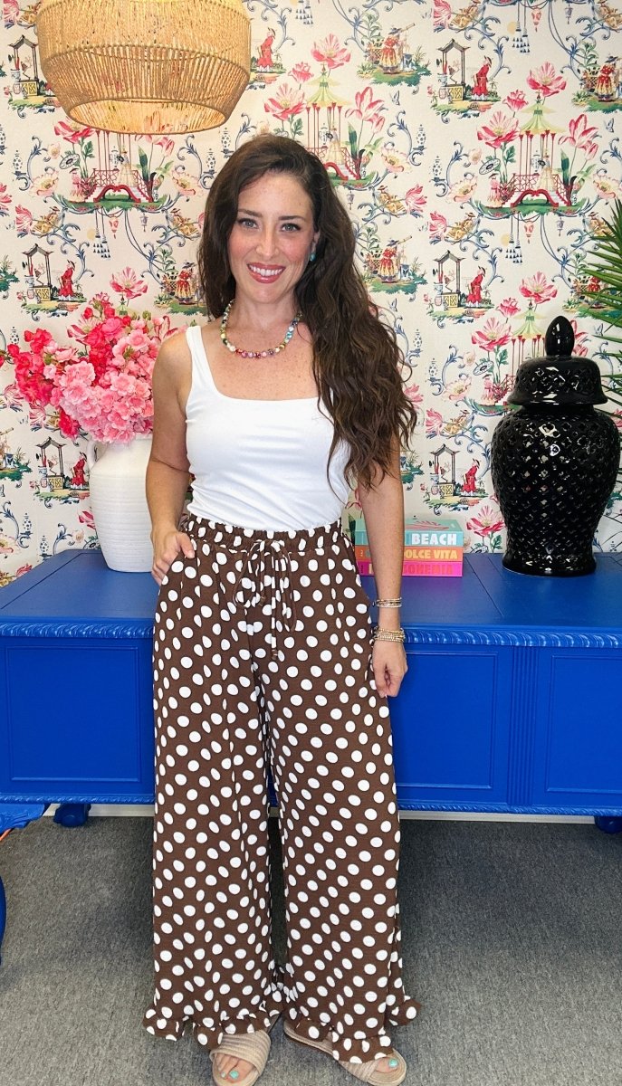 A woman modeling a white tank top and brown wide-leg pants with white polka dots, standing in front of a blue cabinet and floral wallpaper.