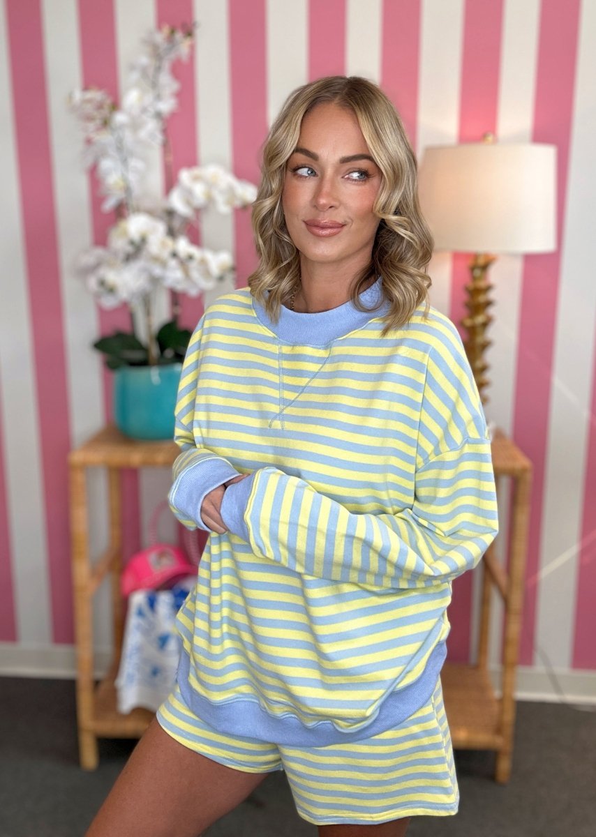 A woman modeling a pastel yellow and light blue striped lounge set, consisting of a long-sleeve sweatshirt and matching shorts, in a room with pink and white striped wallpaper.