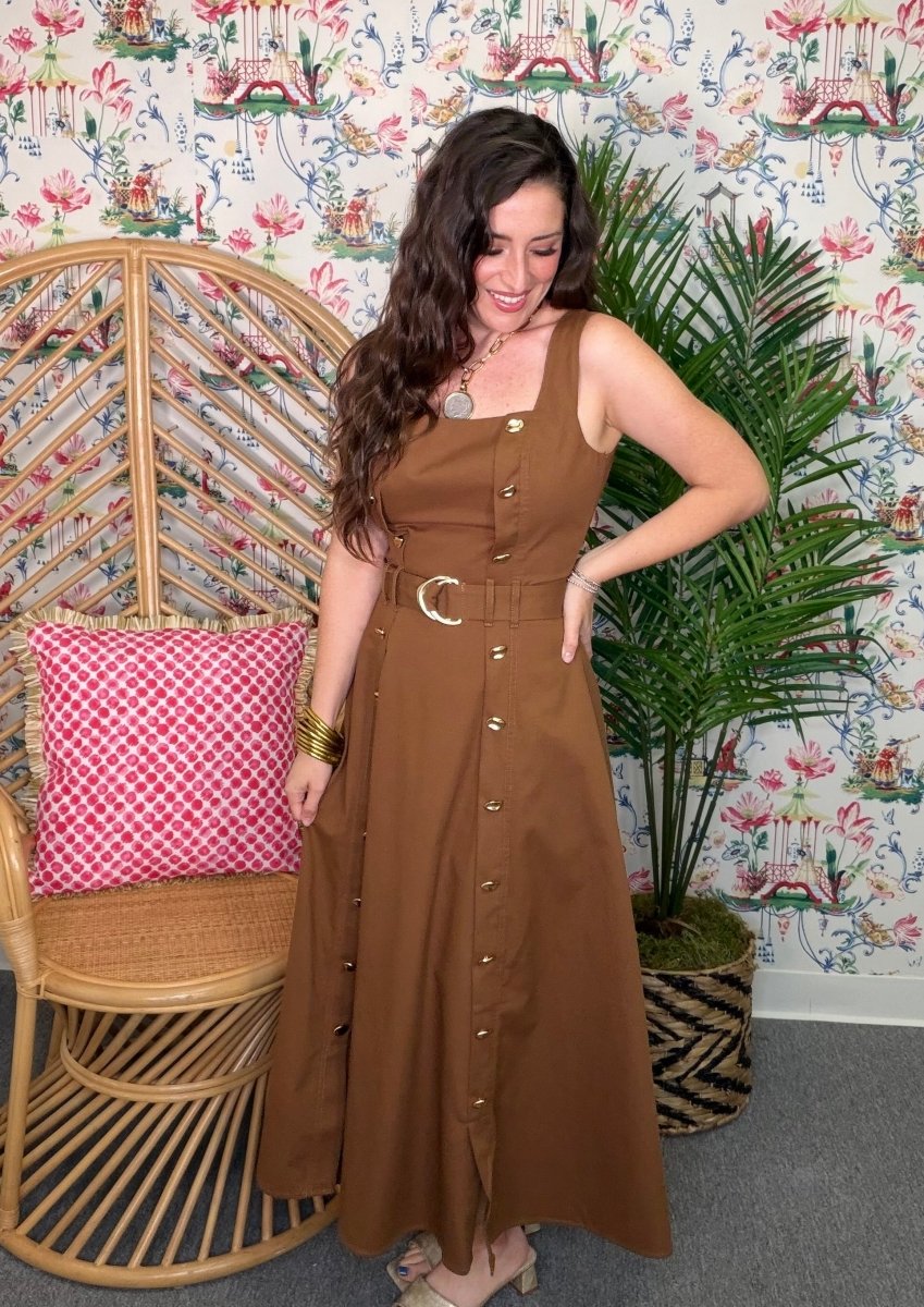 Woman in a brown dress standing in a room with floral wallpaper, a wicker chair, and a plant.