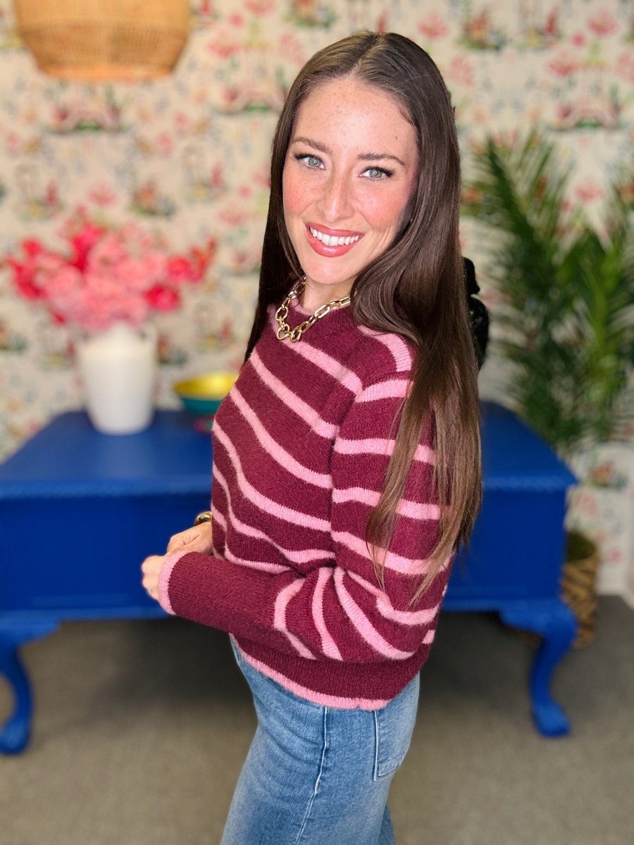 A woman with long brown hair smiling and looking over her shoulder, wearing a burgundy and pink striped knit sweater and blue jeans, standing in a decorated room with floral wallpaper, a blue bench, and plants.