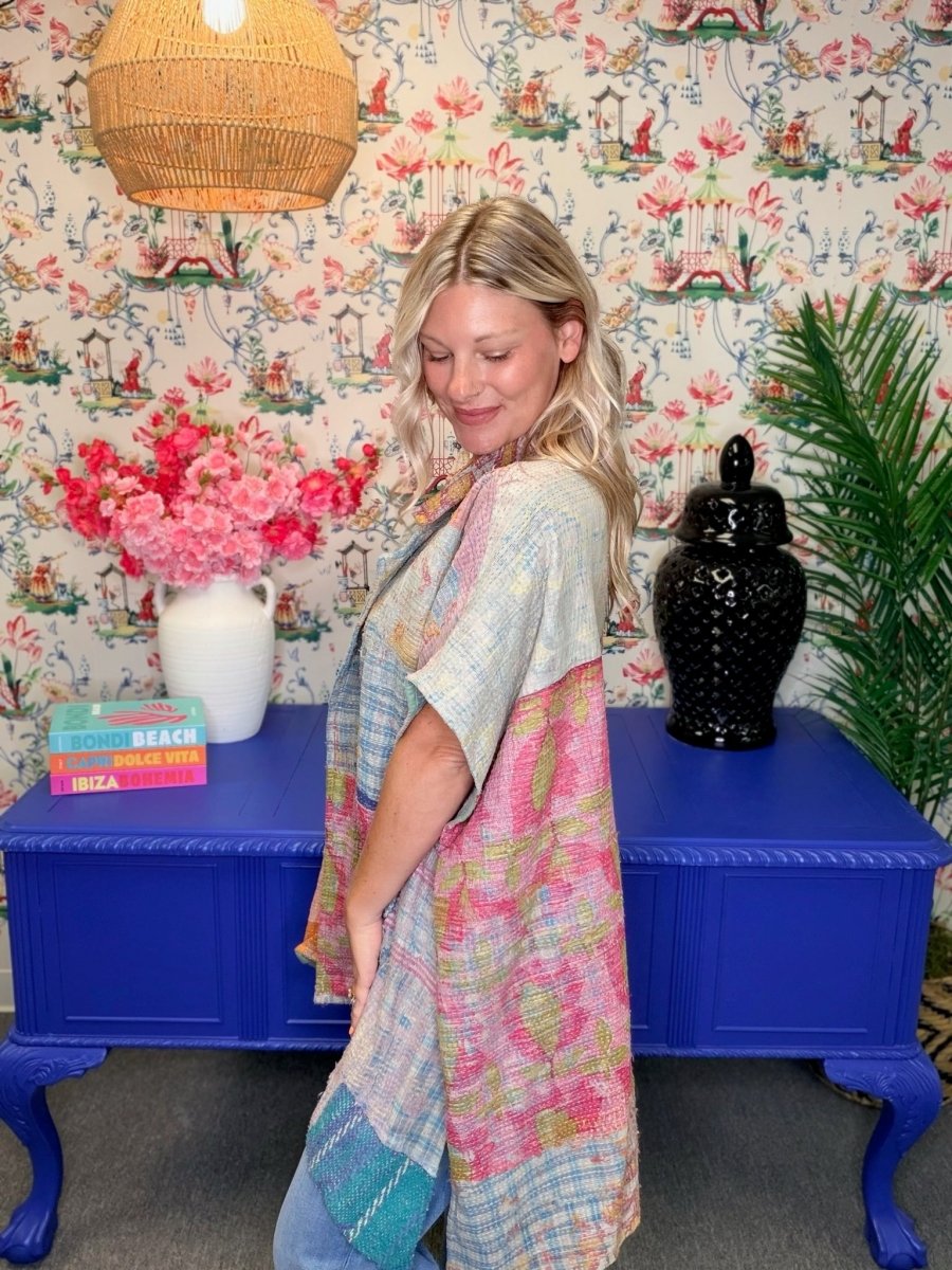 A woman wearing a colorful patchwork tunic top with pink, blue, and green patterns, standing in front of a blue console table decorated with a vase of pink flowers and a black ceramic jar, set against a floral wallpaper background.