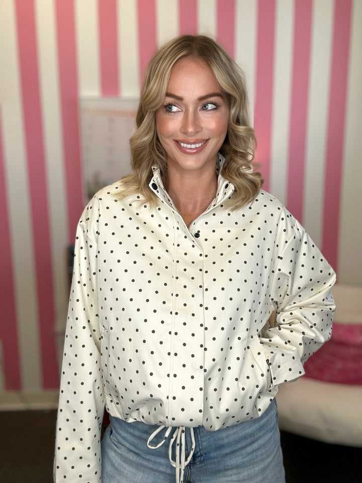 A woman with blonde hair wearing a cream-colored long-sleeve button-up shirt with black polka dots, paired with light blue jeans. She is standing in front of a pink and white striped wall.