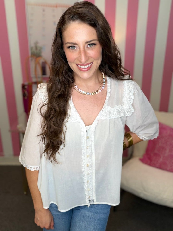Woman wearing a white blouse with lace details in a room with striped wallpaper.