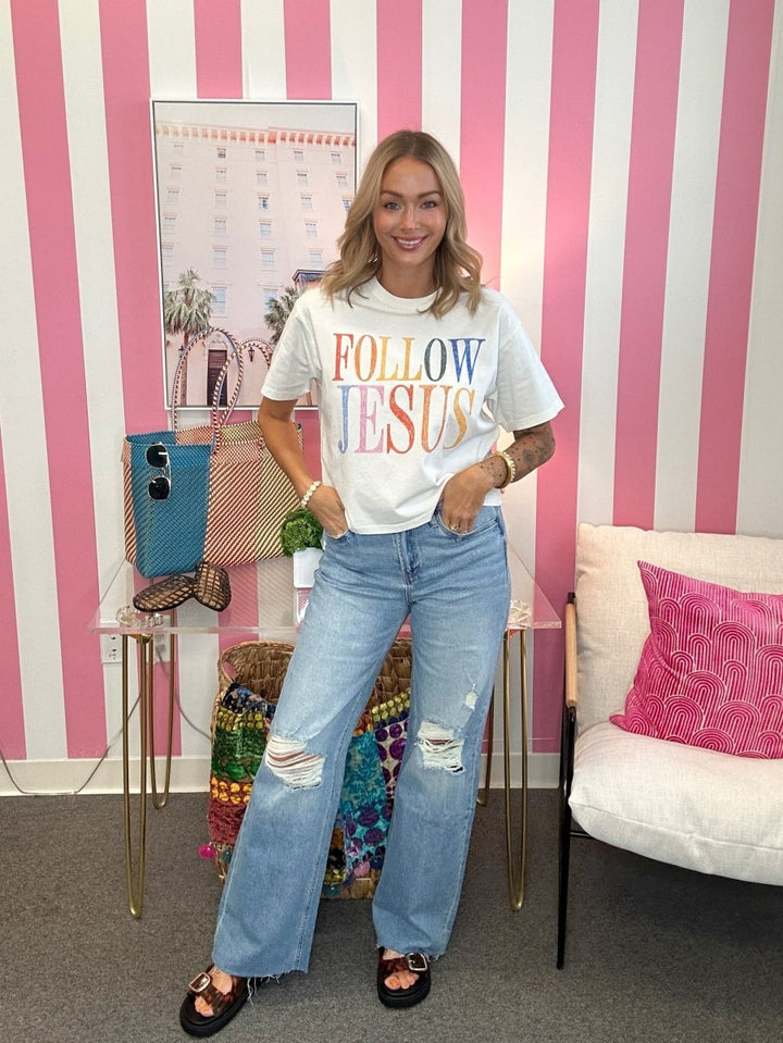 A woman stands in a room with pink and white striped walls, wearing a white graphic T-shirt that reads 'FOLLOW JESUS' in multicolored letters, light-wash distressed jeans, and black sandals.