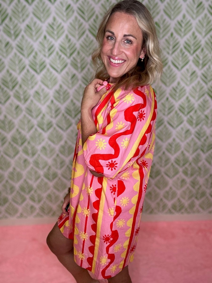 A woman smiling and posing in a pink, red, and yellow patterned kaftan dress against a green and white patterned wallpaper background.