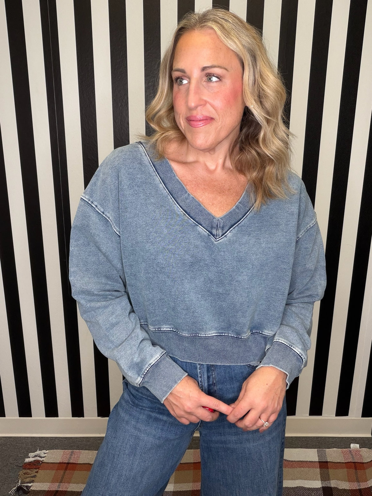 A woman wearing a faded blue denim long-sleeve V-neck crop top and blue jeans, standing in front of a black and white striped wall.