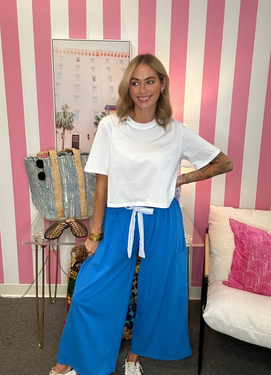 A woman posing in a white short-sleeve crop top and bright blue wide-leg pants with a white drawstring, standing in front of a pink and white striped wall.