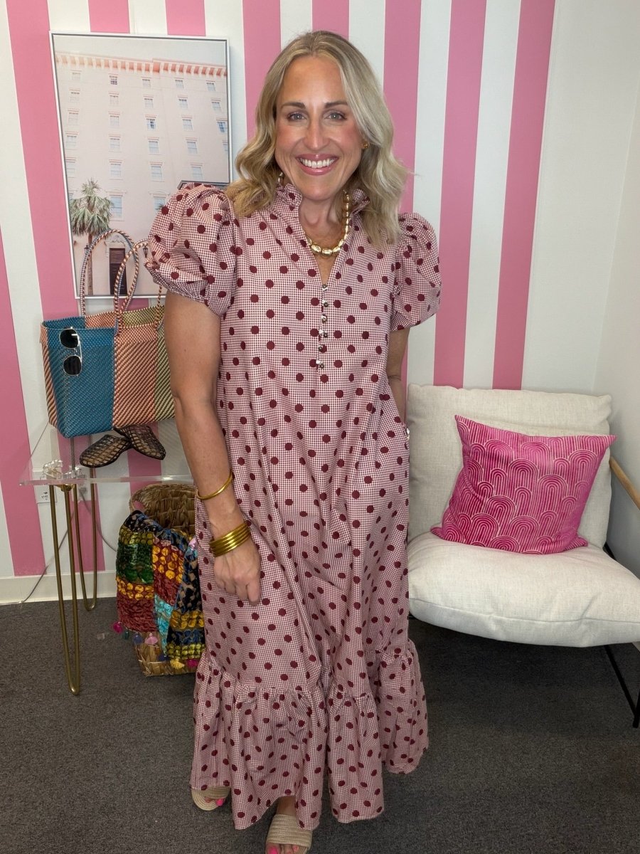 A woman stands indoors wearing a pink maxi dress with dark red polka dots, puff sleeves, and a tiered ruffled hem. The background features pink and white vertical stripes, a white chair with a pink pillow, and a glass side table with handbags.