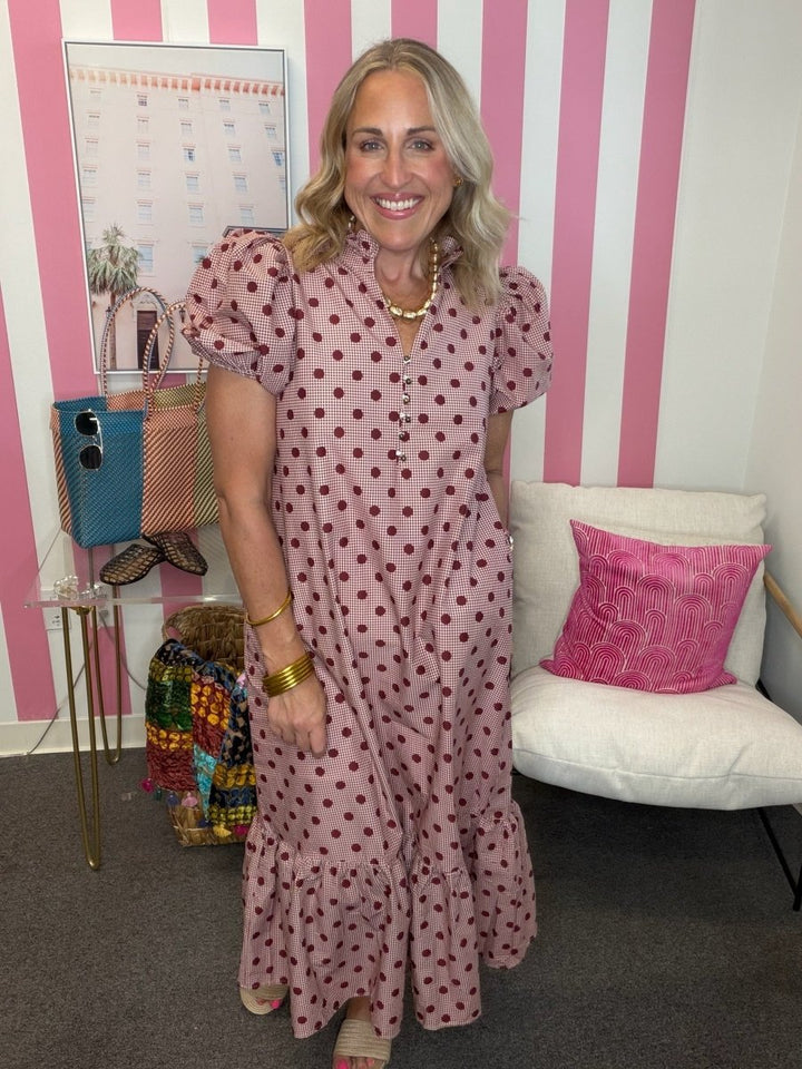 A woman stands indoors wearing a pink maxi dress with dark red polka dots, puff sleeves, and a tiered ruffled hem. The background features pink and white vertical stripes, a white chair with a pink pillow, and a glass side table with handbags.