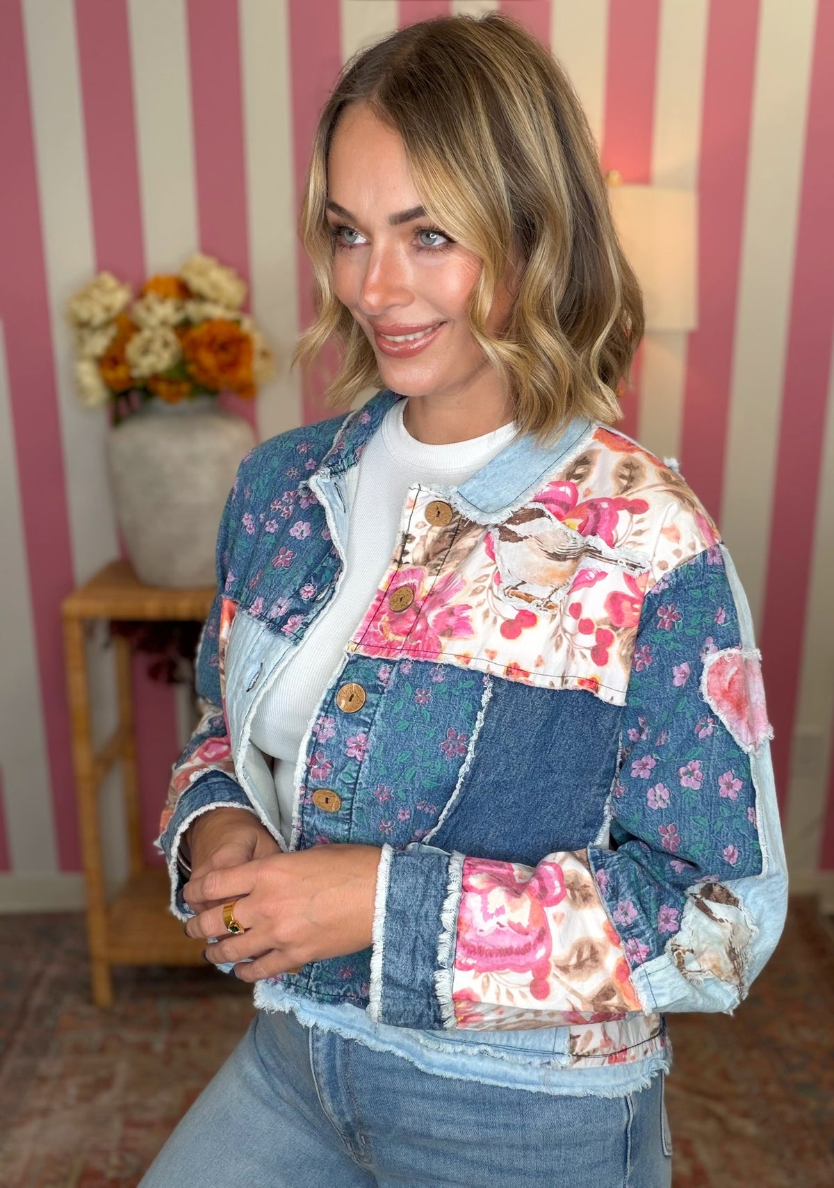 A woman wearing a colorful patchwork denim jacket with floral and teddy bear prints, layered over a white t-shirt and blue jeans, standing in front of a pink and white striped wall.