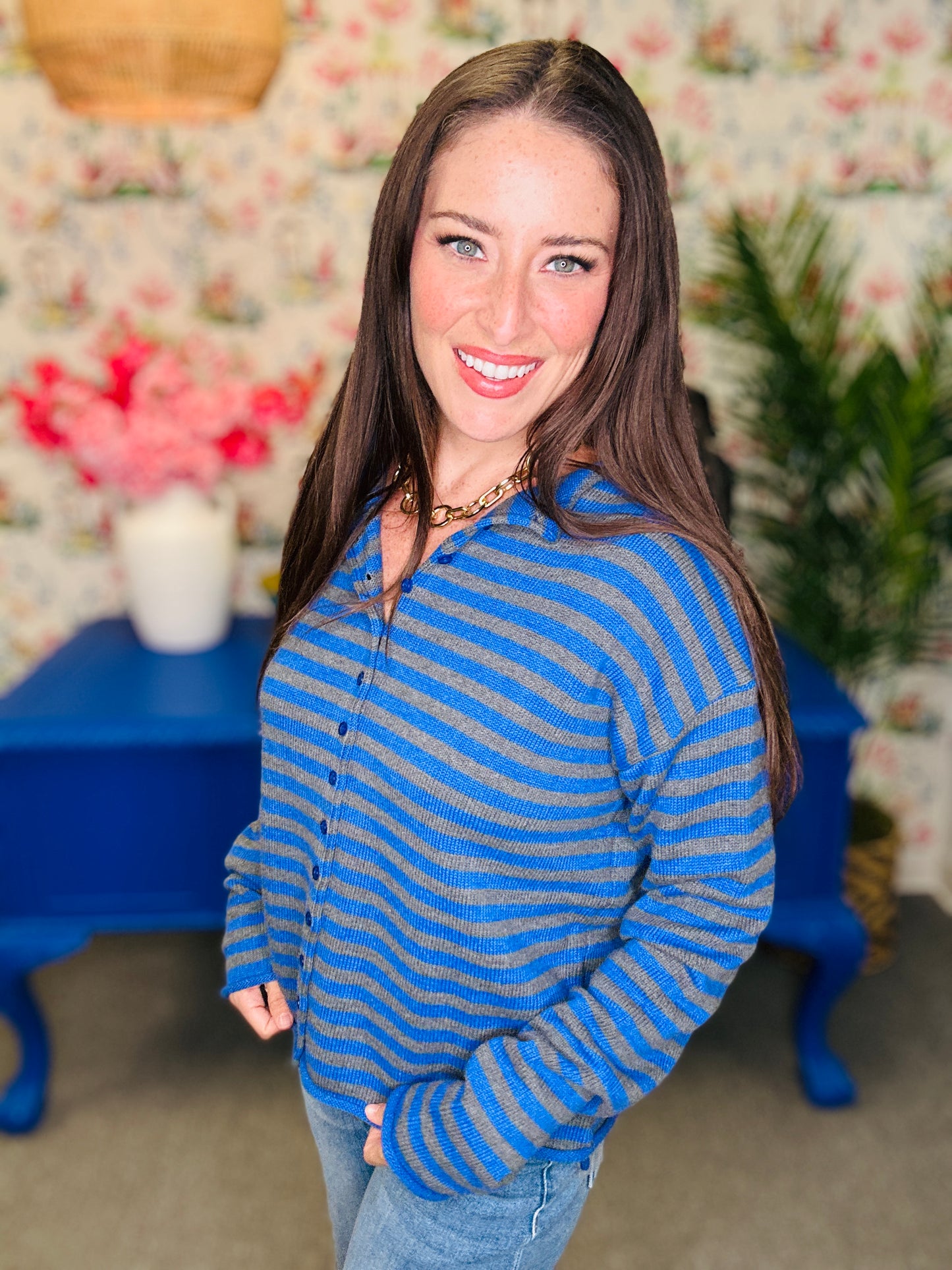 A woman with long brown hair smiling and wearing a blue and gray striped knit cardigan sweater, standing in a decorated room with floral wallpaper, a blue table, and plants.