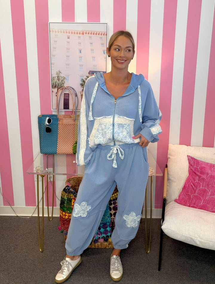 A woman modeling a light blue zip-up hoodie and matching jogger pants set with white lace details, standing in front of a pink and white striped wall.