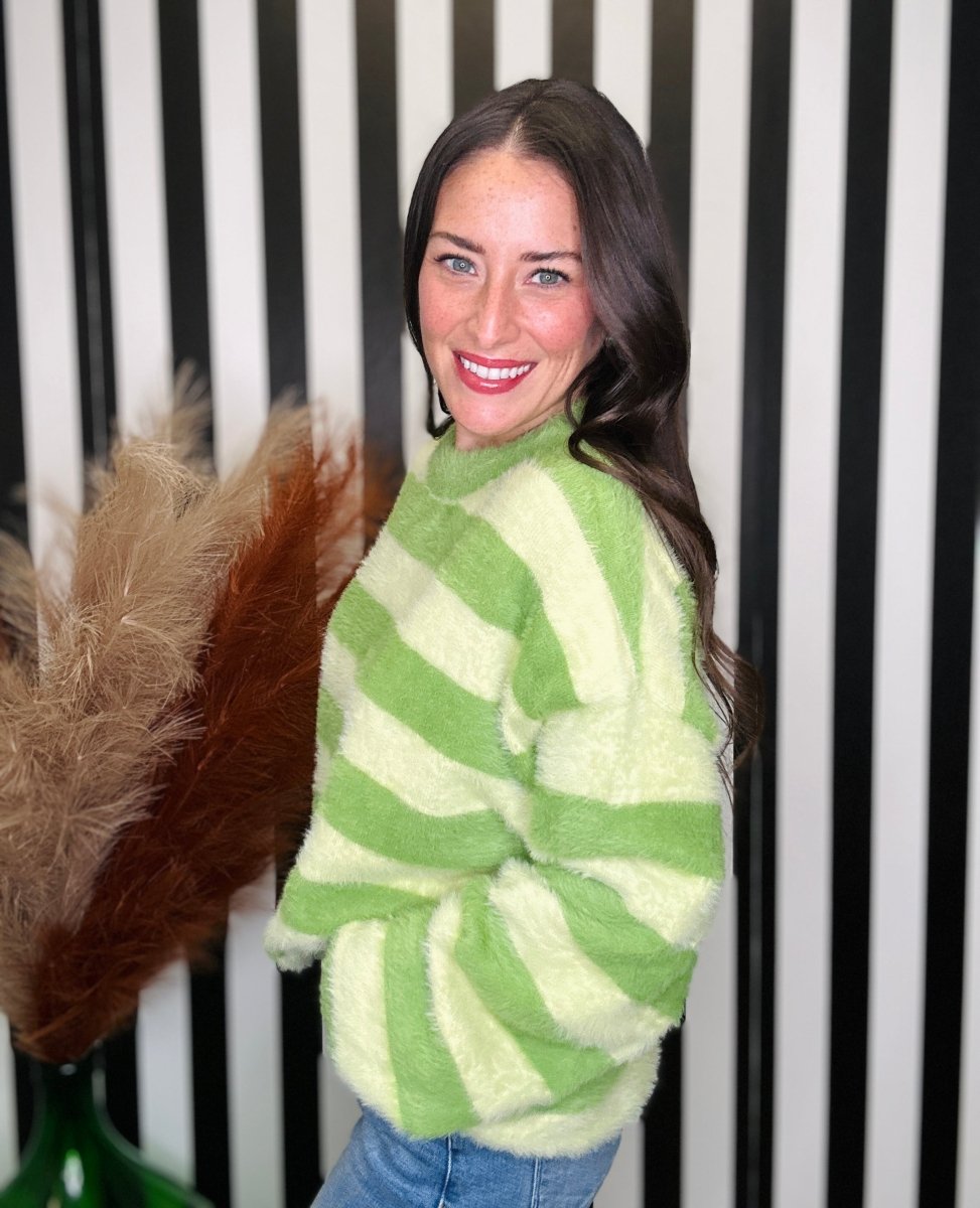 A woman wearing a fuzzy, oversized knit sweater with horizontal lime green and cream stripes, paired with blue jeans, standing in front of a black and white striped wall.