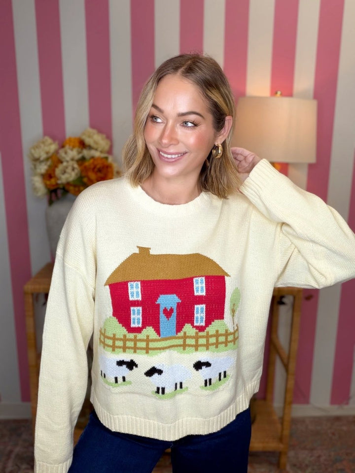 A woman wearing a cream-colored crewneck sweater with a colorful knit design of a red house, blue door, fence, and sheep below it, posed in front of a pink and white striped wall.