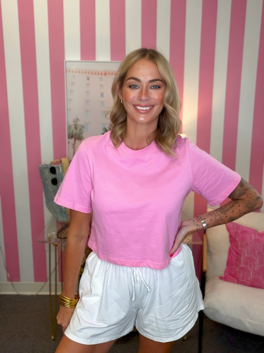 A woman posing in a bright pink short-sleeve crewneck t-shirt and white shorts, standing in front of a pink and white striped wall.