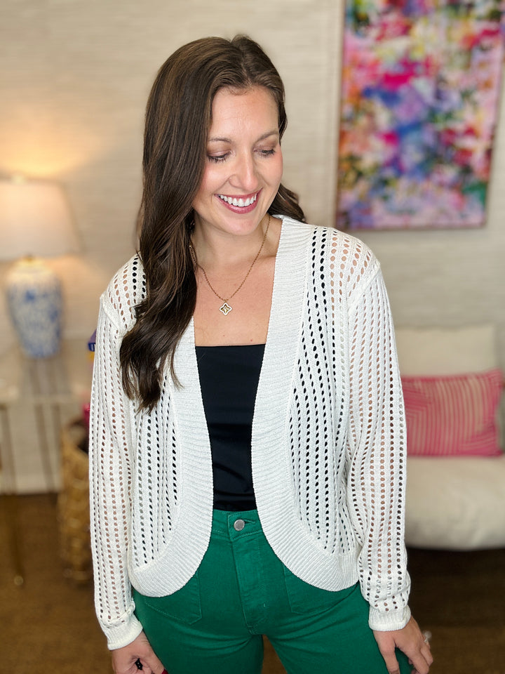 Woman wearing a white open-knit cardigan over a black top and green pants in a living room setting.
