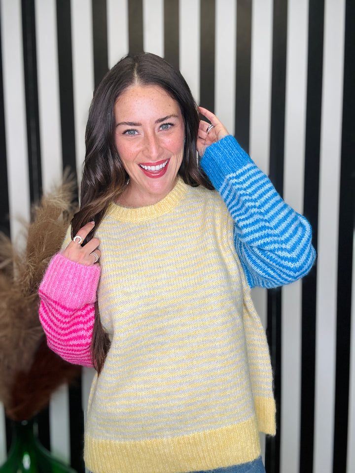 A woman modeling a chunky knit sweater with a yellow body and contrasting sleeves. One sleeve is bright pink with horizontal stripes, and the other is blue with horizontal stripes.