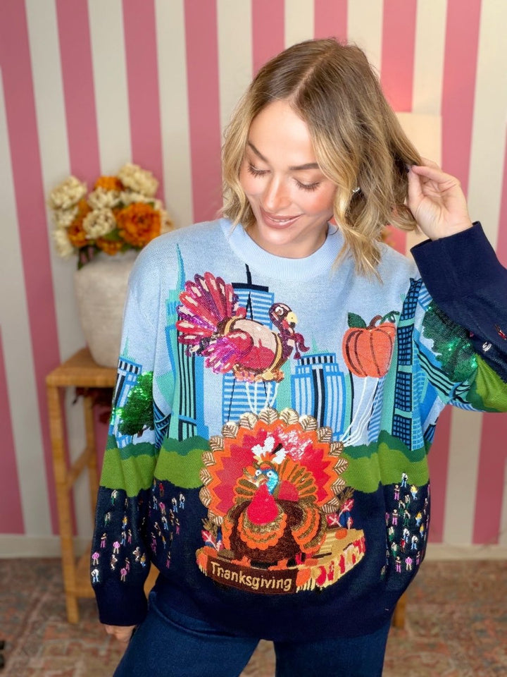 A woman wearing a long-sleeve Thanksgiving-themed knit sweater with a large turkey, pumpkins, and cityscape design, posed in front of a pink and white striped wall.