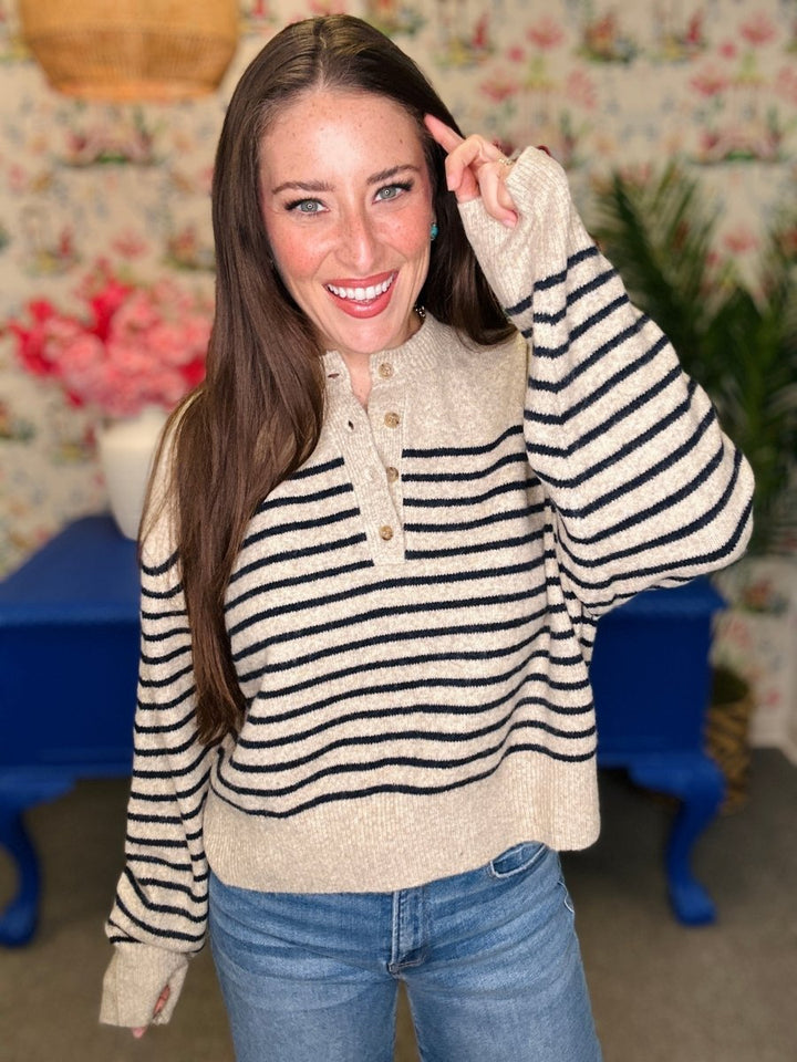 A woman wearing a cream knit sweater with black horizontal stripes and a buttoned henley neckline, paired with blue jeans, standing in a decorated room with floral wallpaper and blue furniture.