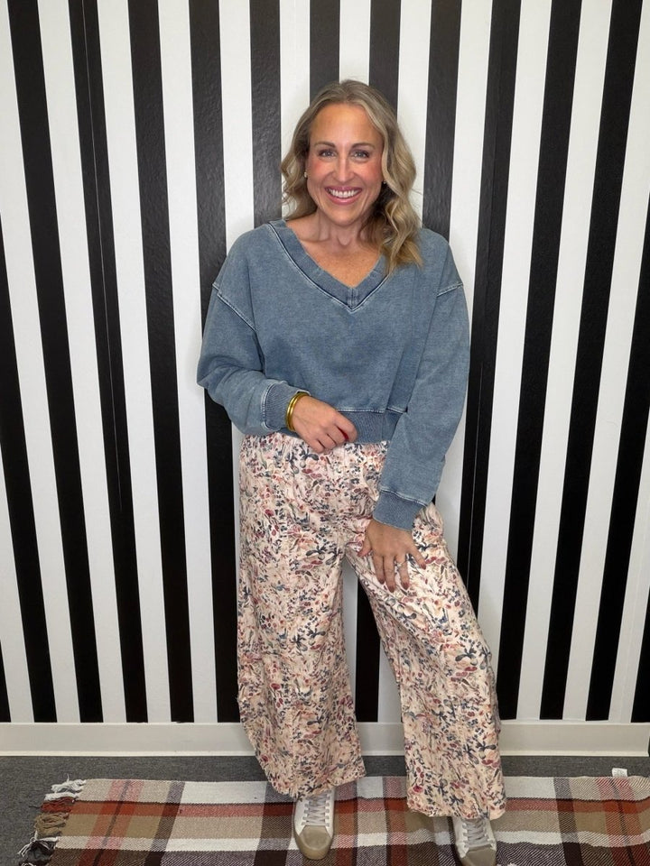 A woman stands in front of a black and white striped wall wearing a blue-gray cropped V-neck sweatshirt and wide-leg floral pants with white sneakers.
