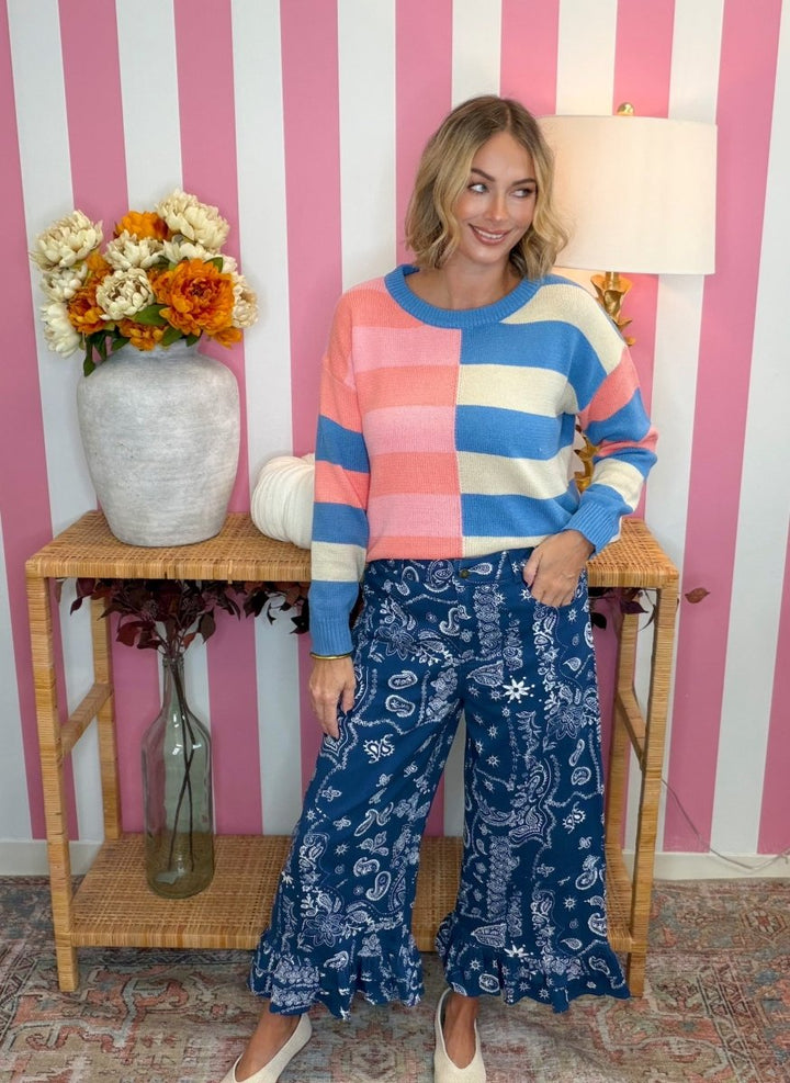 A woman modeling a colorful striped knit sweater and blue paisley wide-leg pants, standing in front of a pink and white striped wall with a side table and flowers.