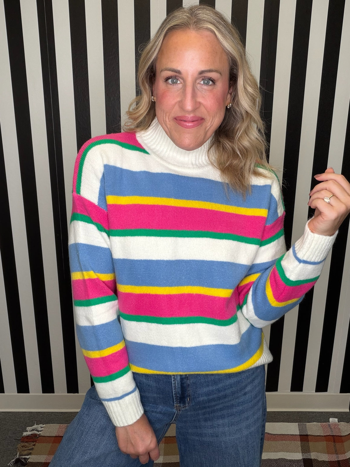 A woman wearing a colorful striped turtleneck sweater with blue, pink, green, yellow, and white horizontal stripes, paired with blue jeans, posed in front of a black and white striped wall.