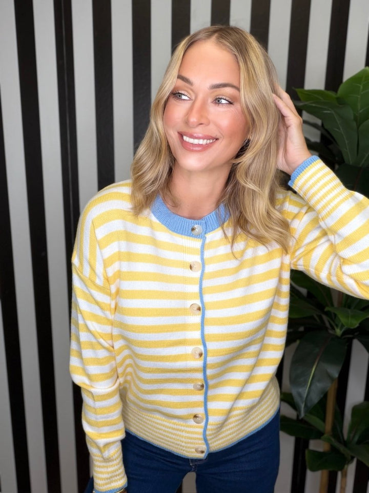 A woman wearing a yellow and white striped knit cardigan with light blue trim, paired with dark blue jeans, standing in front of a black and white striped wall with a green plant in the background.
