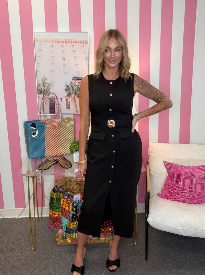 A woman stands indoors wearing a sleeveless black midi dress with a front button placket, waist belt, and front slit. The room has pink-and-white striped walls, a small table with handbags and a colorful quilt.