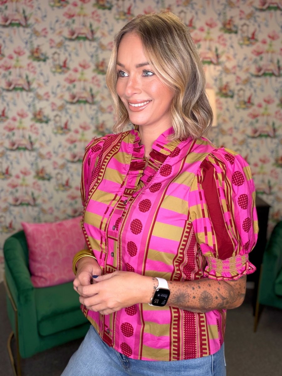A woman wearing a vibrant pink and olive green patterned blouse with puff sleeves, standing in front of a floral wallpaper background.