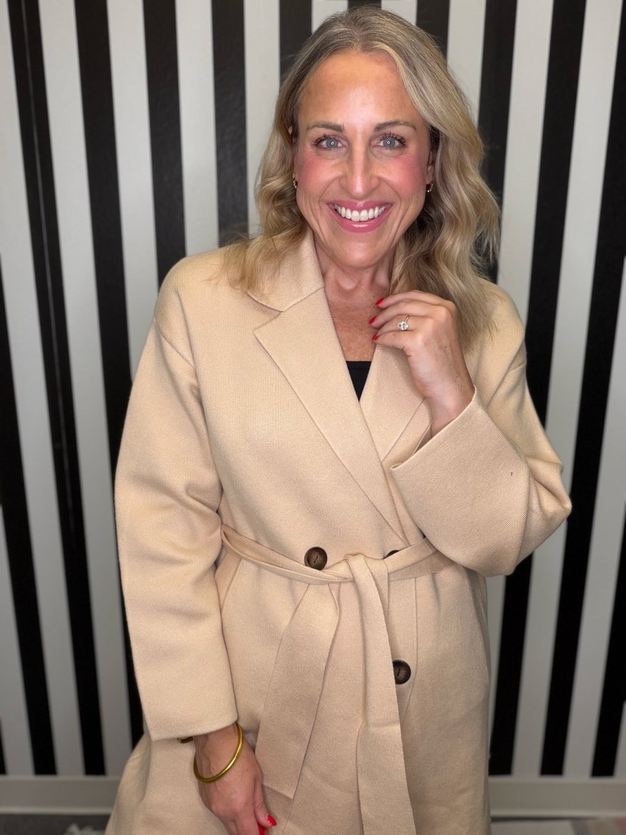 Woman in a brown trench coat standing in front of a black and white striped wall smiling.