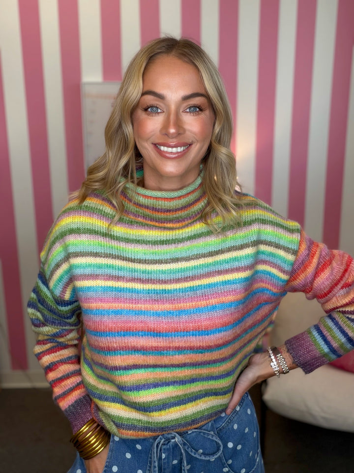 A smiling blonde woman wearing a colorful, rainbow-striped turtleneck sweater and blue polka-dot pants, standing in front of a pink and white striped wall.