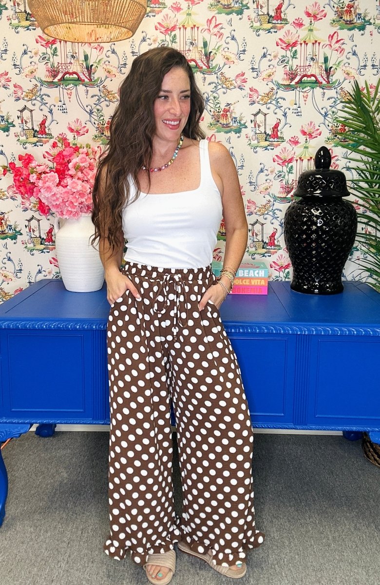 A woman stands in a room wearing brown wide-leg pants with white polka dots, paired with a white tank top. The background features a blue cabinet, floral wallpaper, and decorative items.