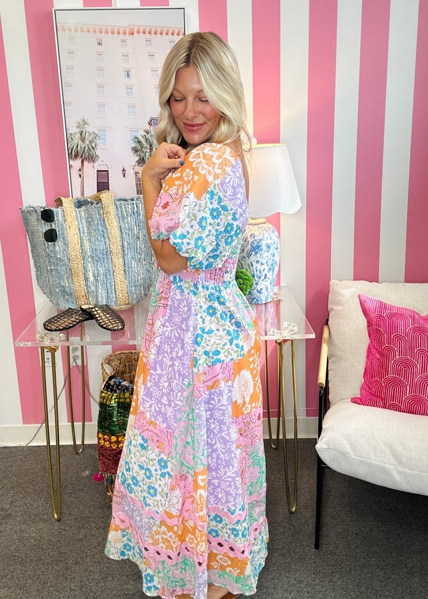 A woman modeling a colorful bohemian-style maxi dress with a floral and abstract print in purple, orange, blue, and pink, standing in a room with pink and white striped walls.