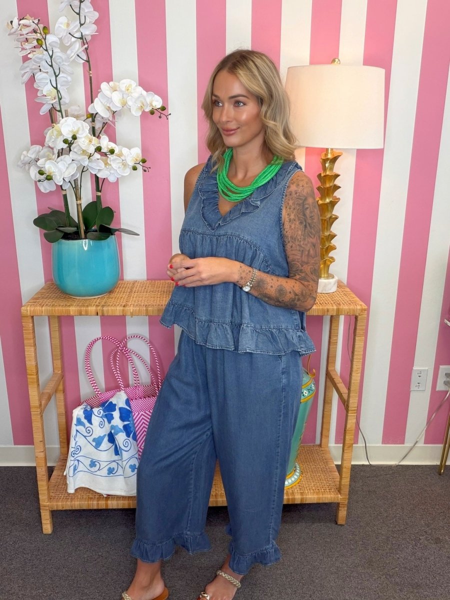 A woman modeling a two-piece denim set consisting of a ruffled sleeveless top and matching wide-leg pants, styled with a bright green necklace, standing in front of a pink and white striped wall.