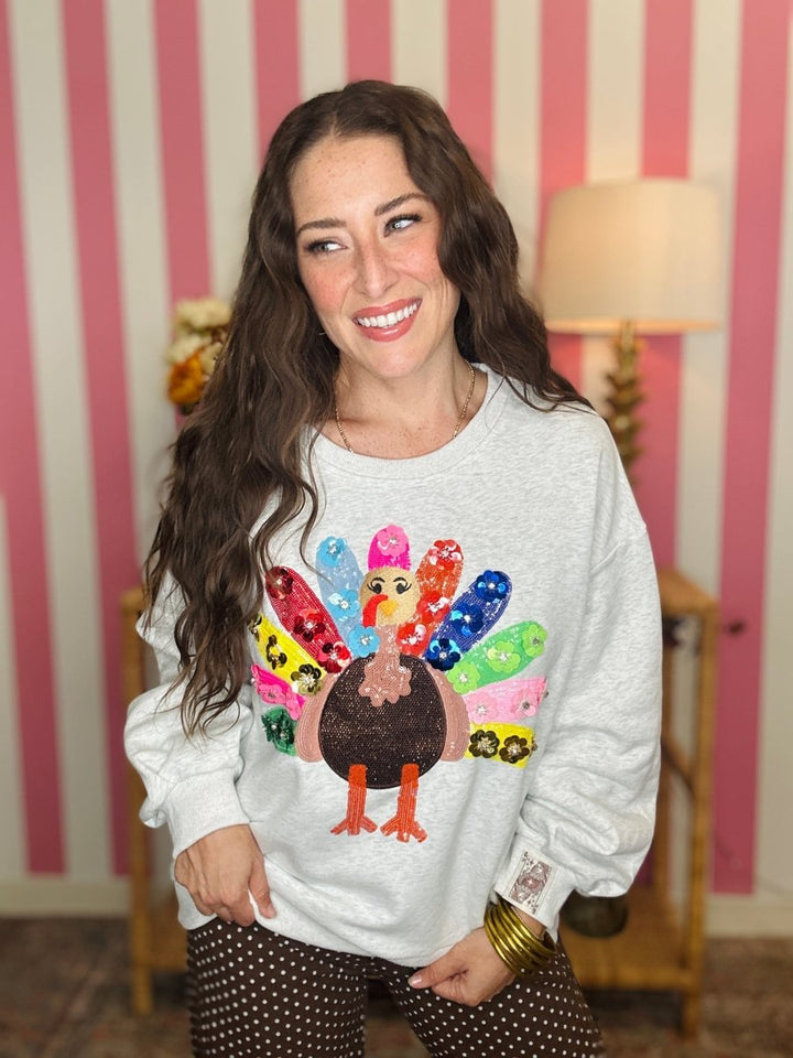 A woman smiling while wearing a light gray crewneck sweatshirt with a colorful sequined turkey graphic on the front, paired with brown polka dot pants, set against a pink and white striped wall background.