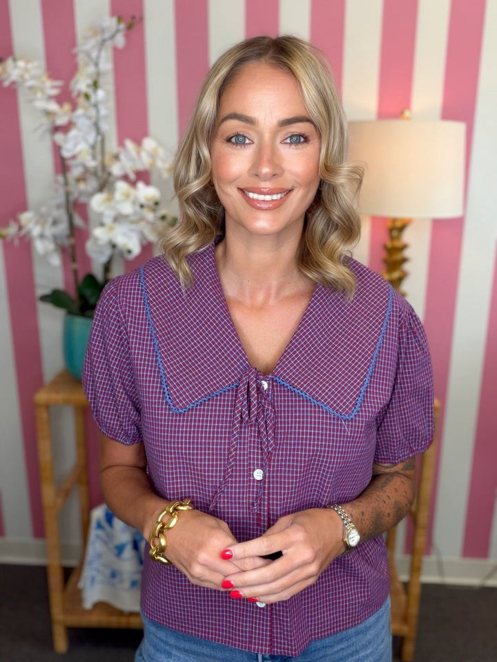 A smiling blonde woman wearing a purple and white gingham short-sleeve blouse with blue piping and a large pointed collar, standing in front of pink and white striped wallpaper.