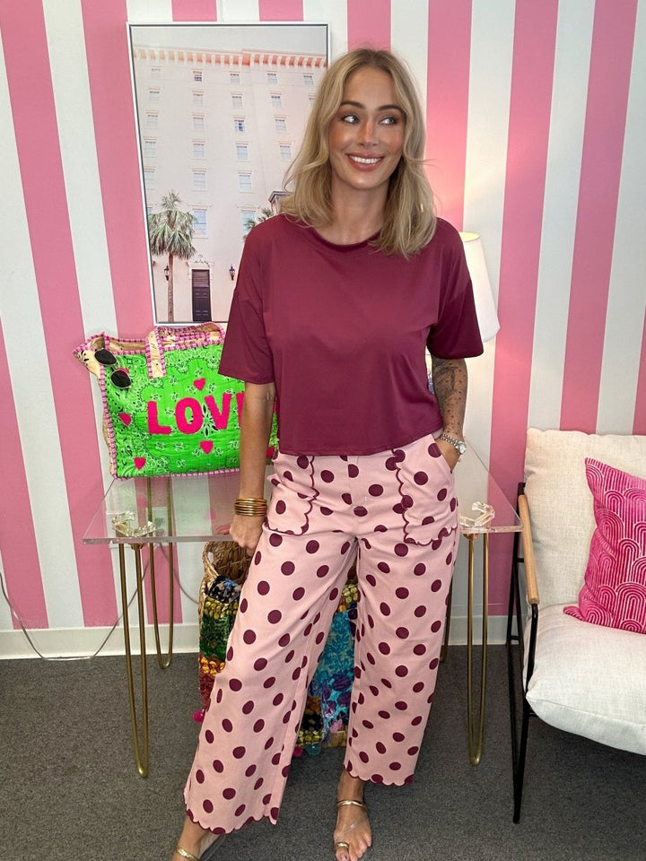 A woman stands in a pink and white striped room wearing a dark burgundy short-sleeve t-shirt and pink wide-leg pants with dark burgundy polka dots. She is standing next to a glass table holding a green and pink 'LOVE' handbag.