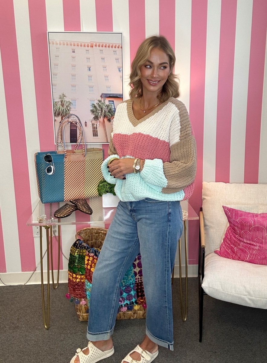 A woman stands in a room with pink and white striped walls, wearing a color-block knit sweater with beige, cream, pink, and light blue sections, paired with blue jeans and white slide sandals.