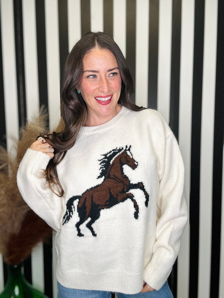 A woman wearing a cream-colored knit sweater with a brown and black embroidered horse design on the front, standing in front of a black and white striped background.