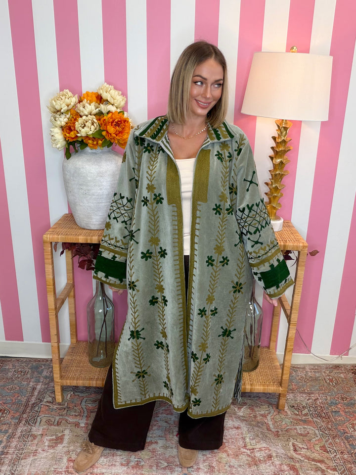 A woman stands in a room with pink and white striped wallpaper, wearing a long sage green embroidered cardigan with olive green trim and dark green geometric patterns. She is standing behind a small wooden side table decorated with a white vase of orange and white flowers and a gold lamp.