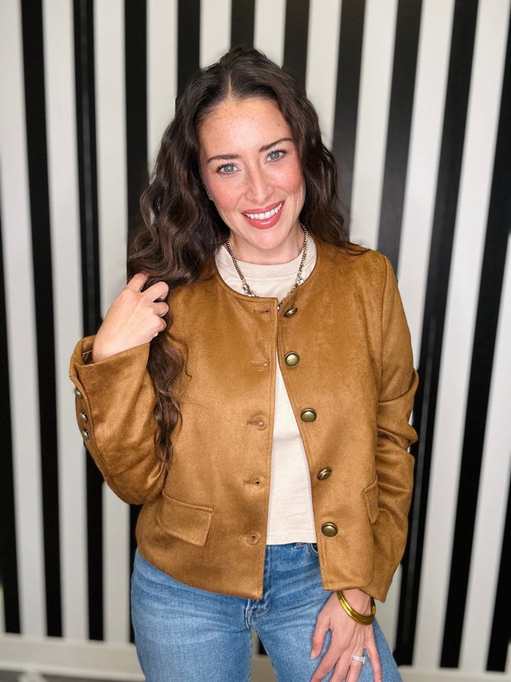 A woman with long wavy brown hair smiling, wearing a cropped tan suede jacket with gold-tone buttons over a cream turtleneck and blue jeans, standing in front of a black and white vertically striped background.