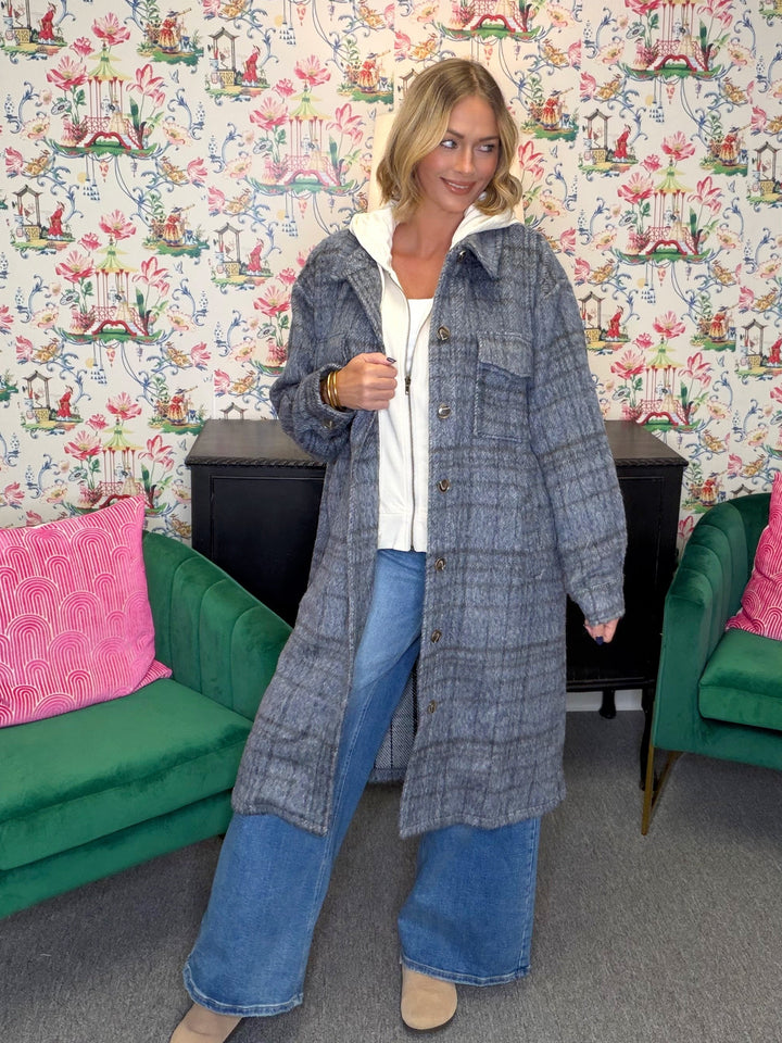 A woman stands in a room with floral wallpaper, green velvet chairs, and a black cabinet, wearing a long gray plaid button-up coat over a white top and wide-leg blue jeans.